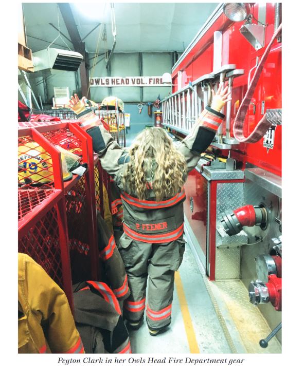 Peyton Clark in her Owls Head Fire Department gear. Peyton is standing back to the camera with hands up in the air. Next to her is a bright red fire engine, and various fire outfits and cubbies on her other side. Photo by Becca Shaw Glaser.